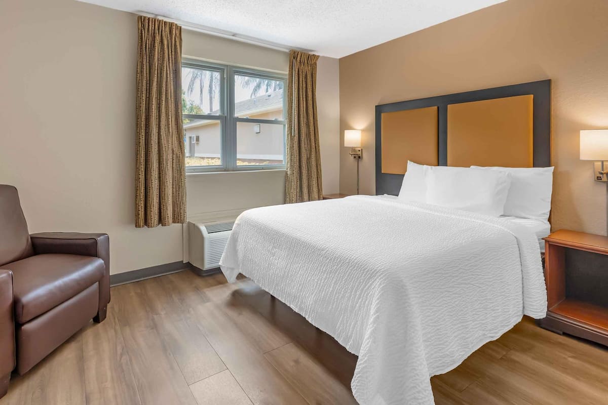 Ein Hotelzimmer mit Bett, Stuhl, einem Fenster mit Vorhängen und einer Wandlampe.