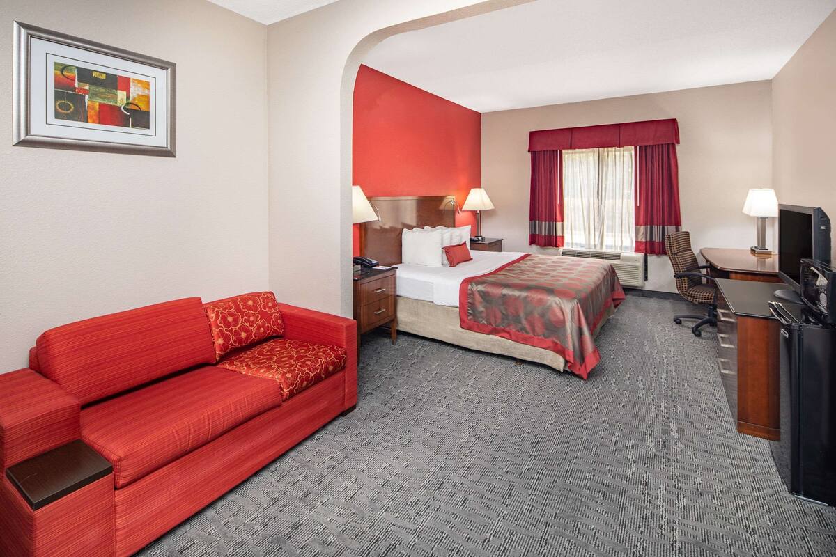 A hotel room with a red sofa, a bed with a red bedspread, a desk with a computer, and a TV.