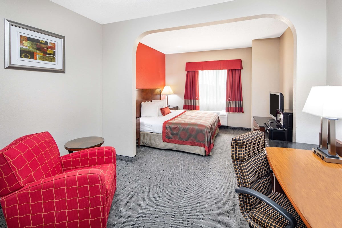 A hotel room with a bed, a red armchair, a small round table, a wooden desk, and a television.