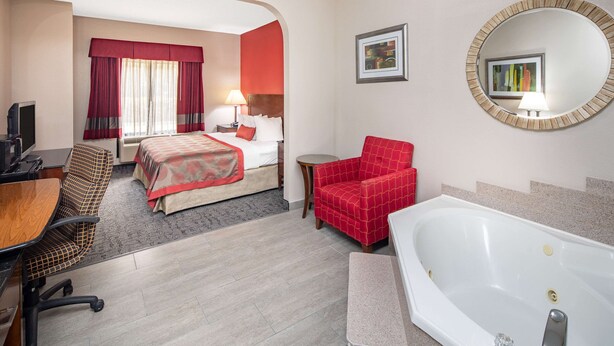 A hotel room with a bed, a desk, a chair, a red armchair, a round mirror, and a bathtub. - Ramada by Wyndham Alpharetta/Atlanta North (Alpharetta)