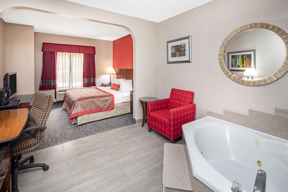 A hotel room with a bed, a desk, a chair, a red armchair, a round mirror, and a bathtub.