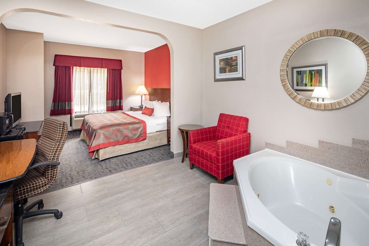 A hotel room with a bed, a red armchair, a desk with a chair, a round mirror, and a bathtub.