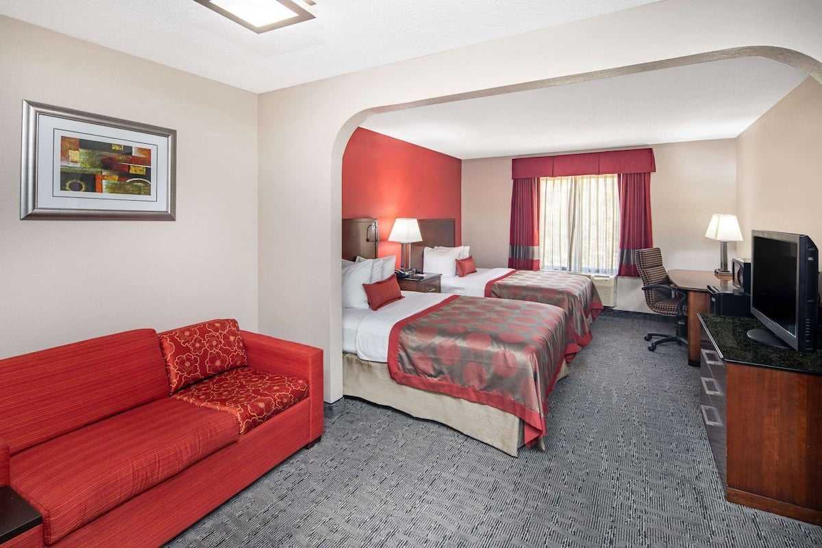 A hotel room with a red sofa, two beds with matching red and beige bedding, a desk with a television, and a framed picture on the wall.