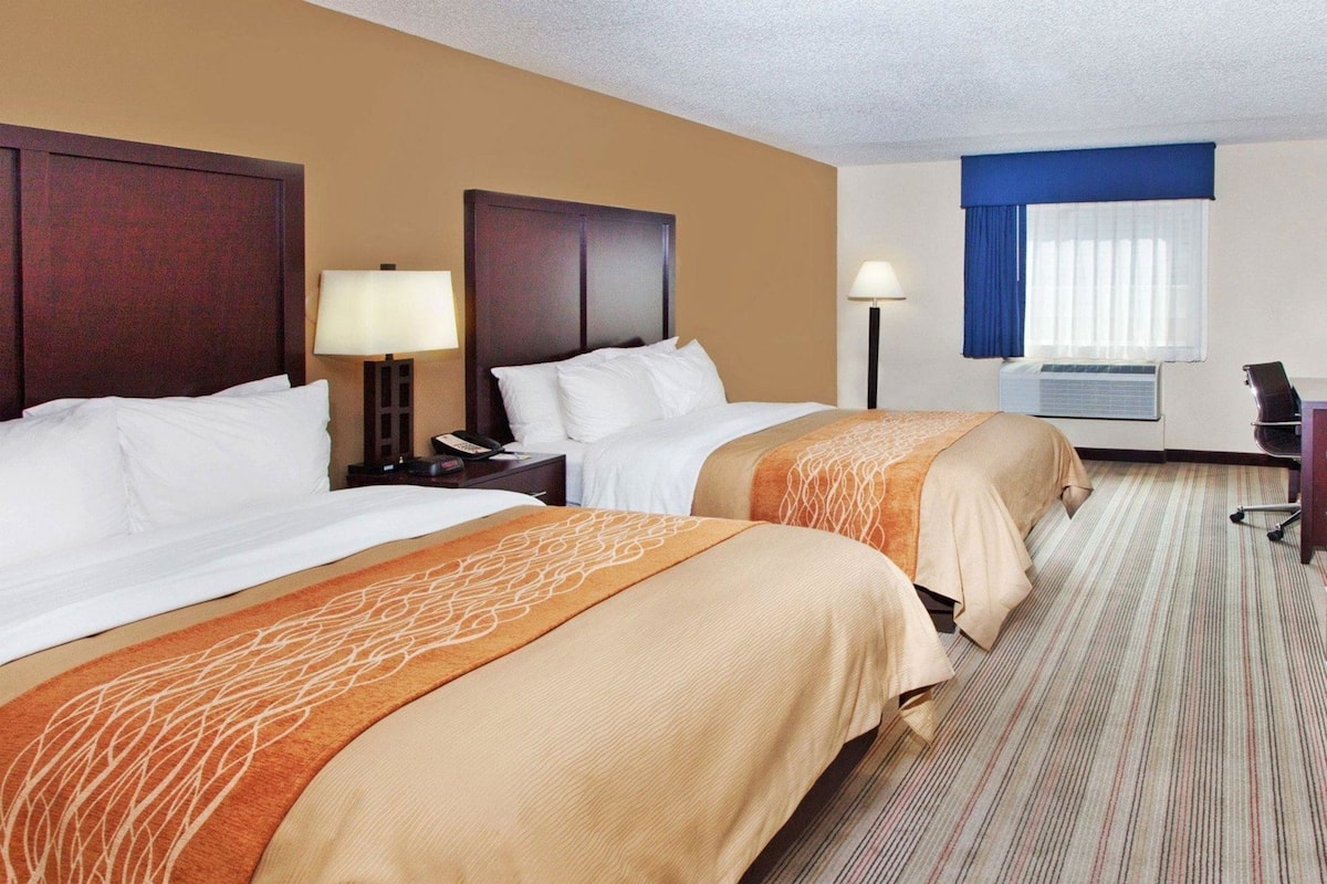 A hotel room with two beds, a desk, a chair, a lamp, and an air conditioning unit.