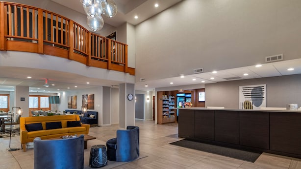 A spacious lobby with a wooden staircase, a reception desk, and a seating area with a yellow sofa and blue ottomans. - Best Western Plus Dalton Inn (Dalton)
