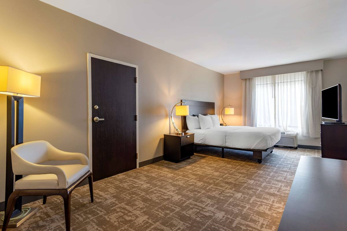 A hotel room with a bed, a chair, a nightstand with a lamp, a television, and a large window with curtains.