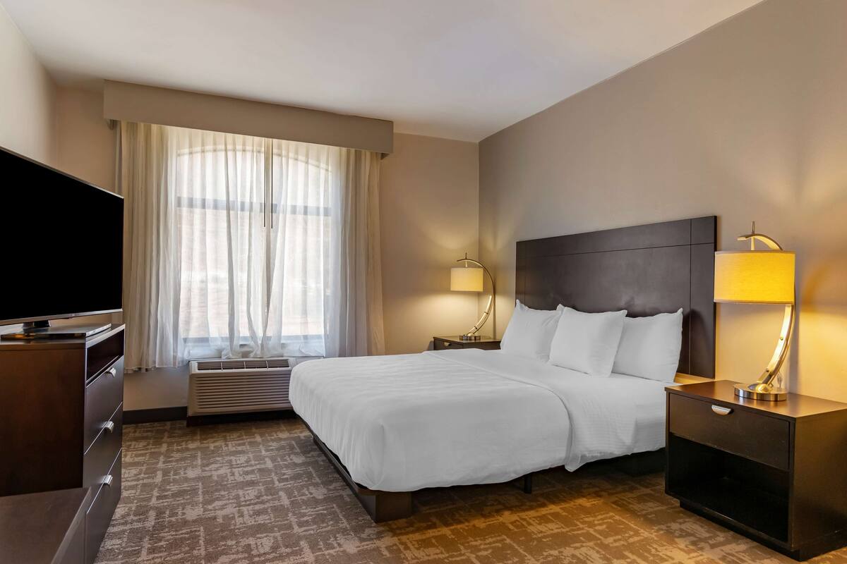 A hotel room with a large bed, a flat-screen TV, a desk with a lamp, and a window with sheer curtains.