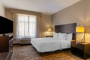 Una habitación de hotel con una cama grande, un televisor de pantalla plana, un escritorio con lámpara y una ventana con cortinas translúcidas.