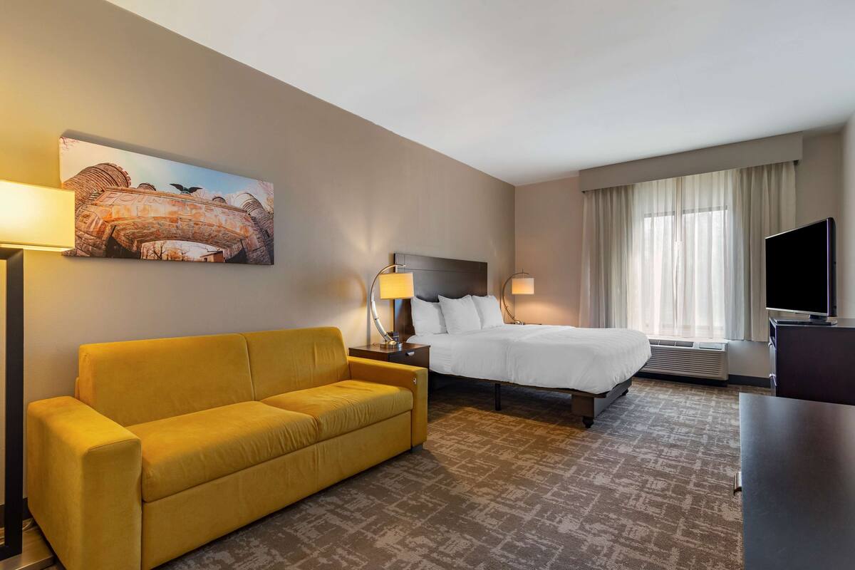 A hotel room with a yellow sofa, a bed, a TV, and a painting on the wall.