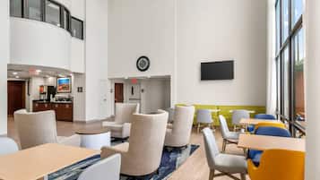 En hotelllobby med sittgrupper, en klocka, en platt-TV och ett kök synligt i bakgrunden.