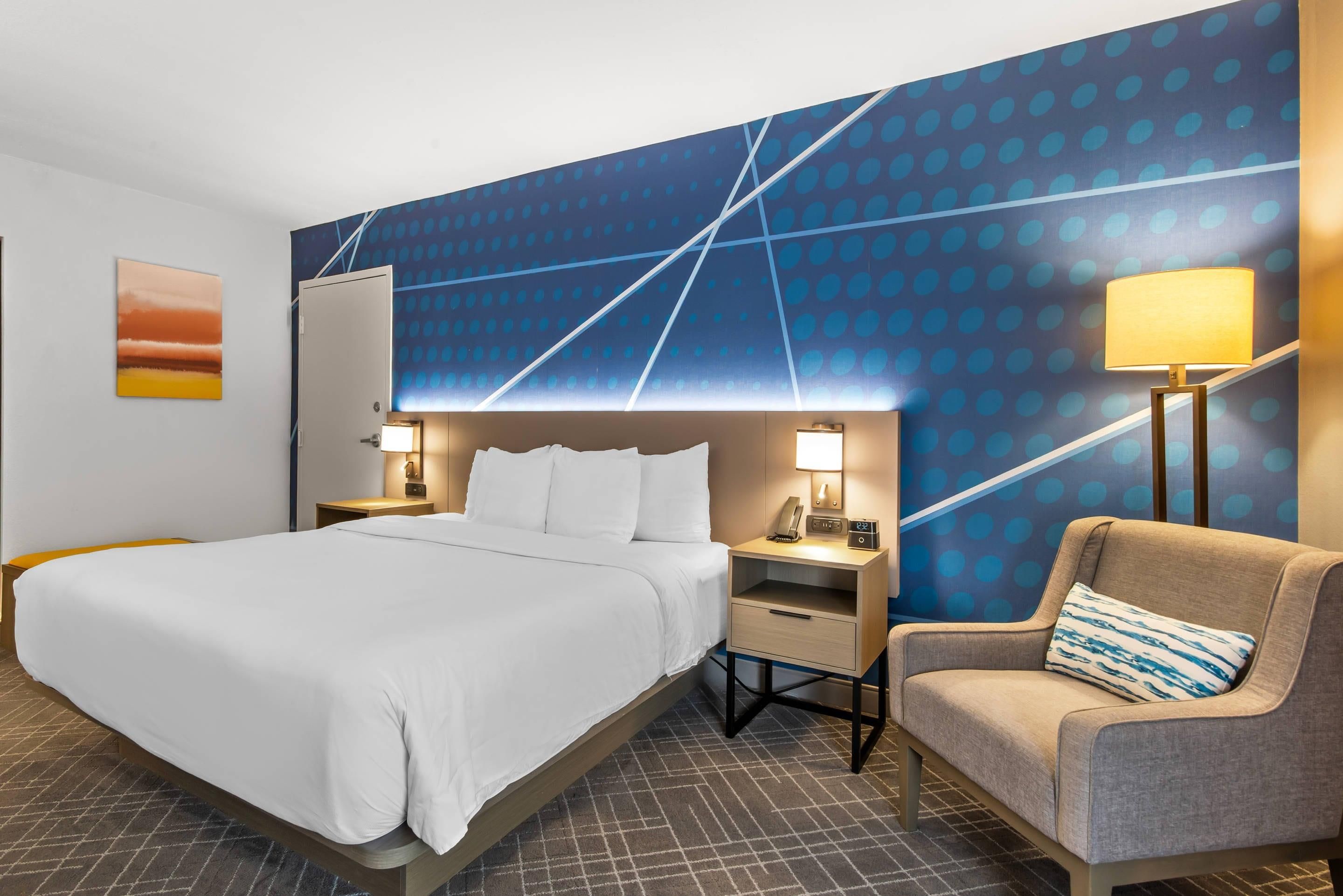 Quarto de hotel com cama, mesa de cabeceira, luminária, cadeira e parede com padrão geométrico.