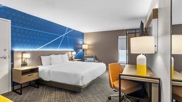 A hotel room with a bed, desk, chair, nightstand, lamp, and a wall with a geometric pattern.
