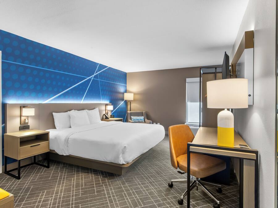 A hotel room with a bed, desk, chair, nightstand, lamp, and a wall with a geometric pattern.