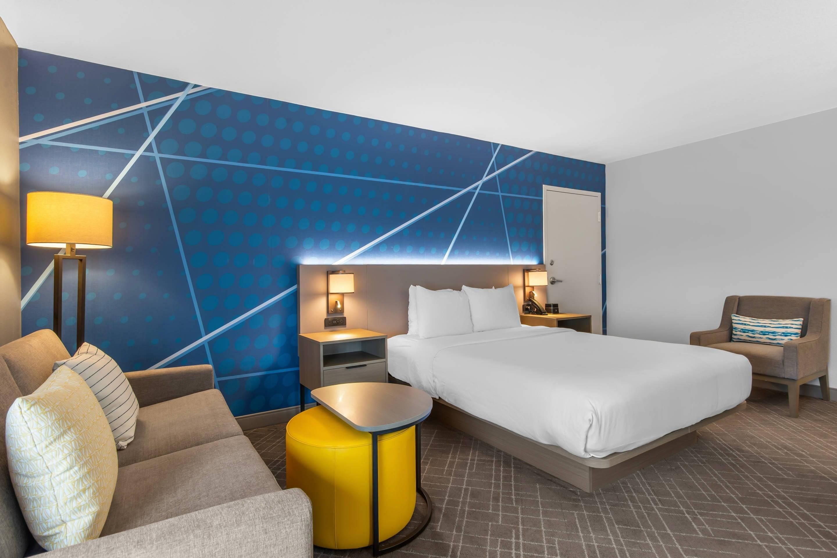 Quarto de hotel com uma cama, um sofá, uma mesinha, uma cadeira, uma mesaninha de cabeceira e uma parede com um padrão geométrico.