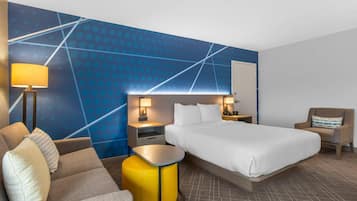 A hotel room with a bed, a sofa, a small table, a chair, a nightstand, and a wall with a geometric pattern.
