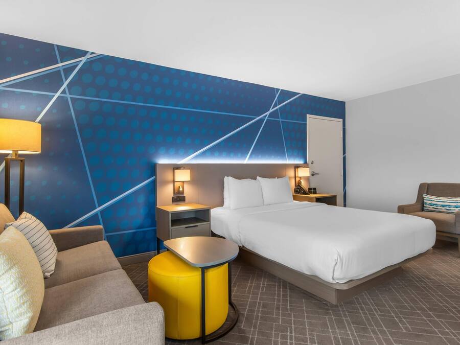 A hotel room with a bed, a sofa, a small table, a chair, a nightstand, and a wall with a geometric pattern.