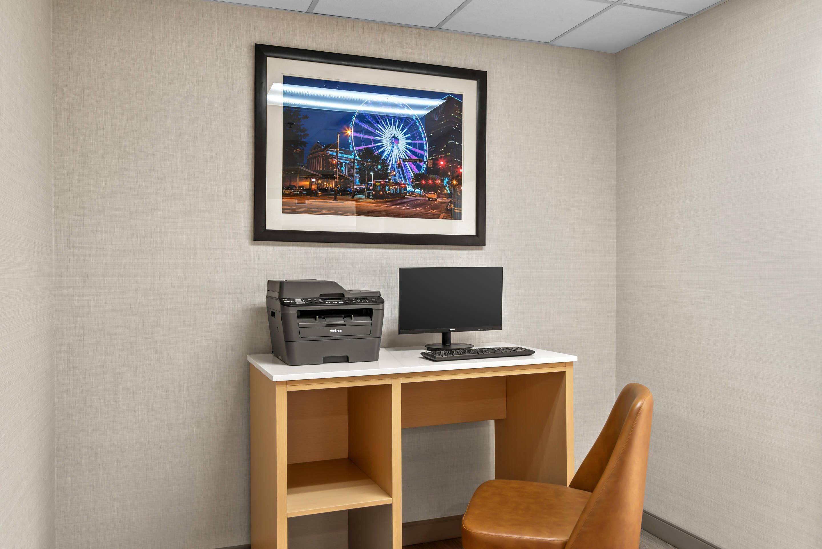 A desk with a printer and monitor, a framed picture of a Ferris wheel, and a brown chair.
