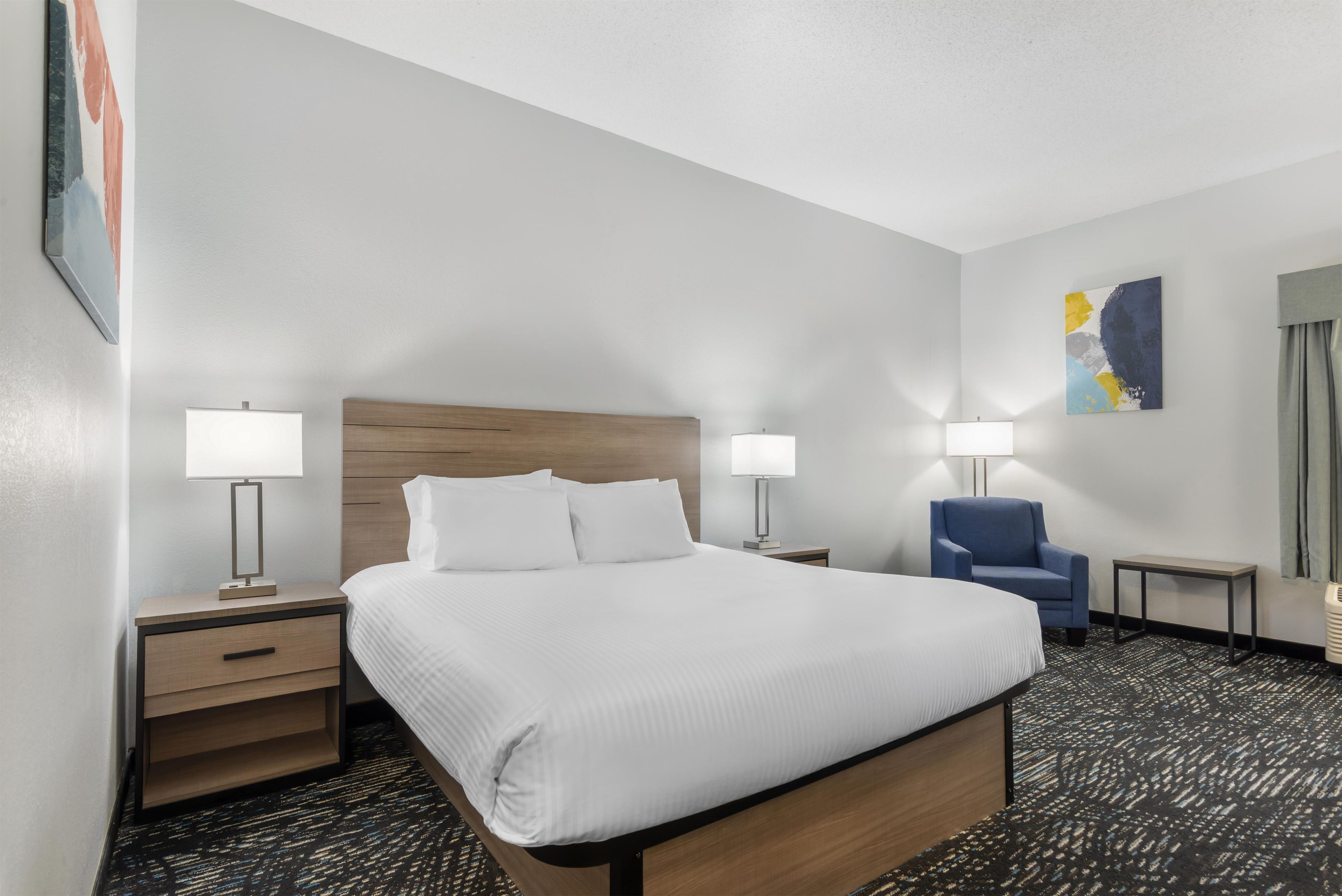 Una habitación de hotel con una cama grande, dos mesitas de noche con lámparas, una silla, una mesita pequeña y un cuadro en la pared.