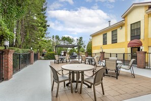 Ein Außenbereich mit Terrassenmöbeln, einer überdachten Sitzgelegenheit und einem Gebäude mit gelben Wänden und Fenstern.