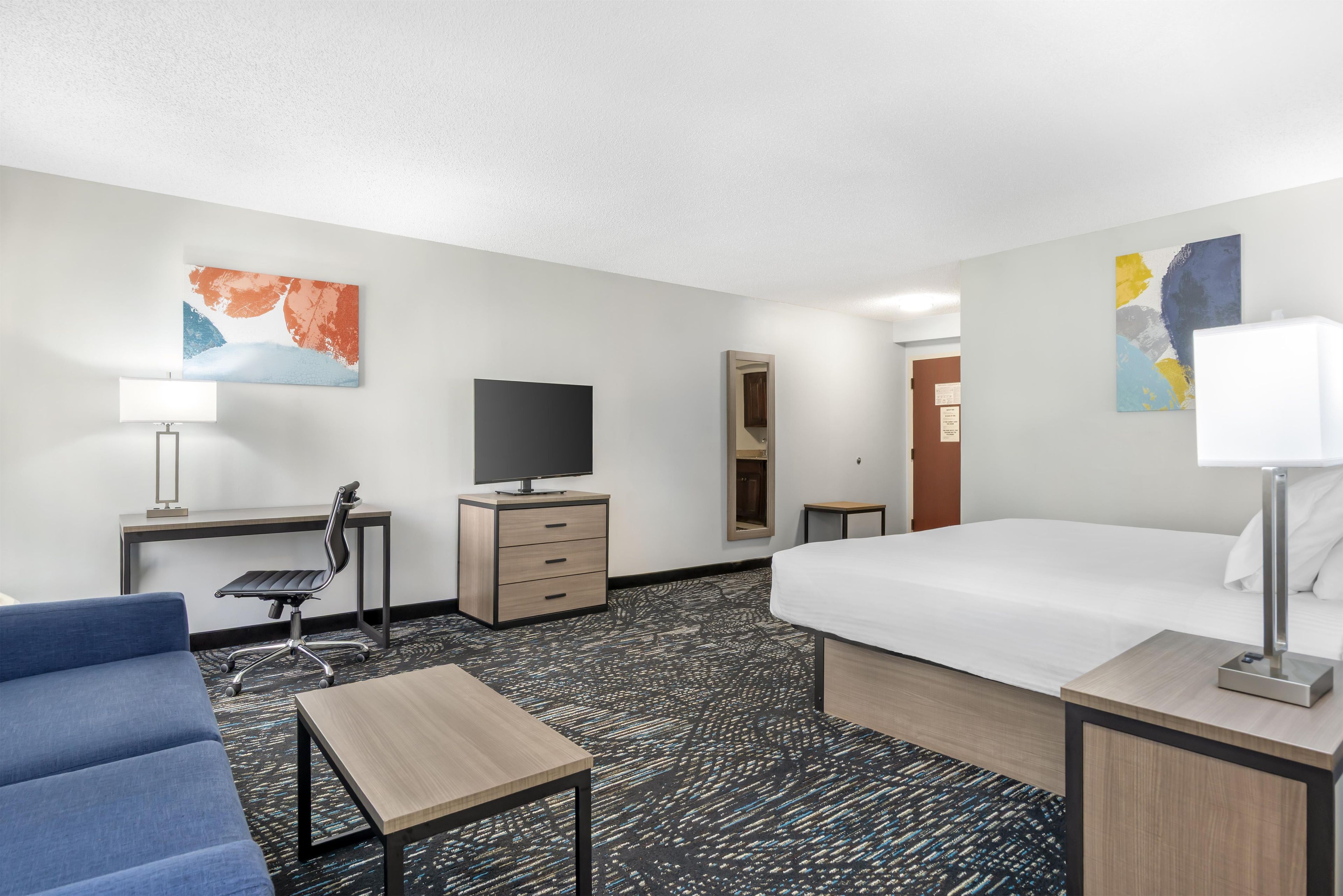 A hotel room with a bed, a desk with a chair, a television, a dresser, and artwork on the walls.