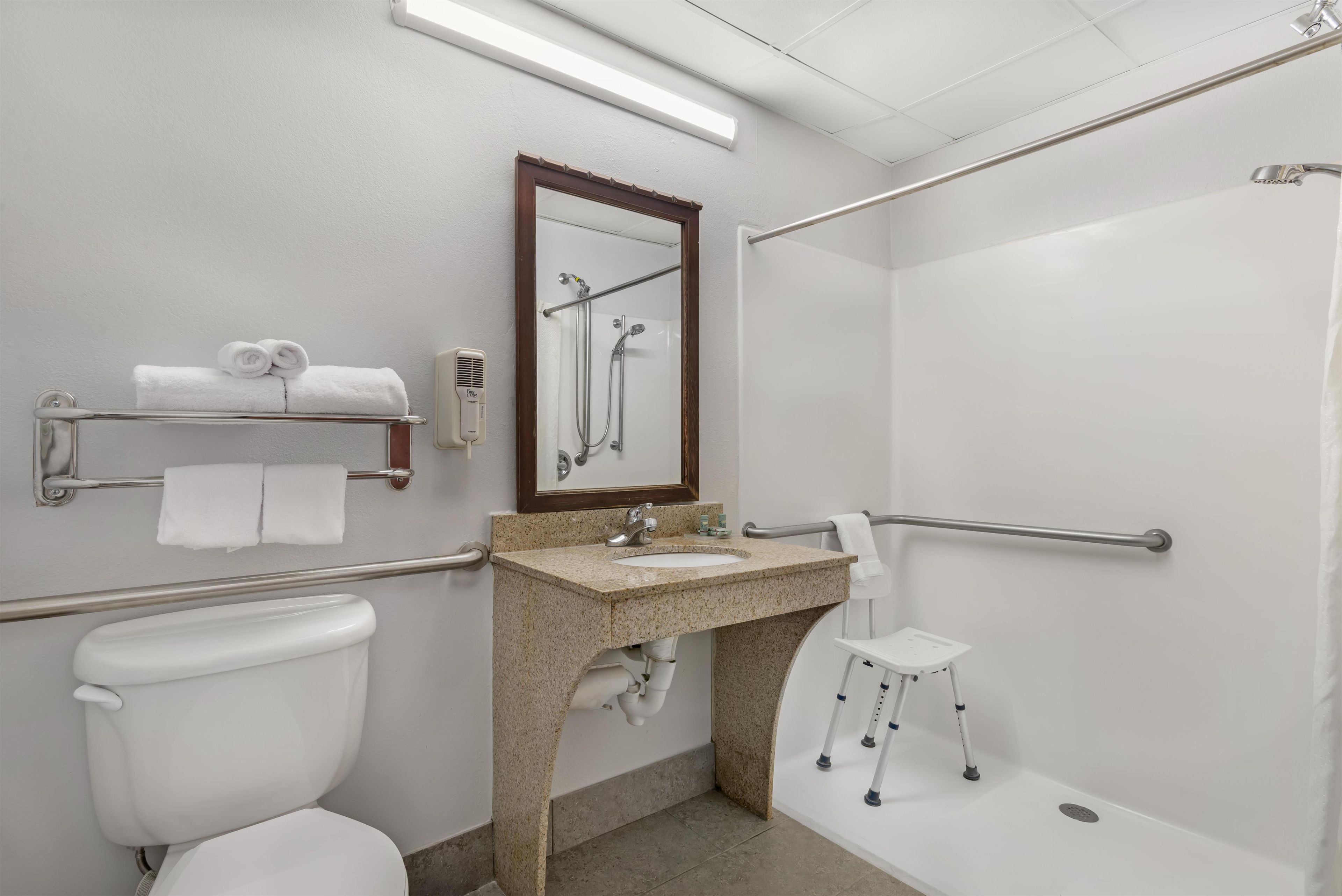 A bathroom with a toilet, sink, mirror, towel rack, shower, and grab bars.