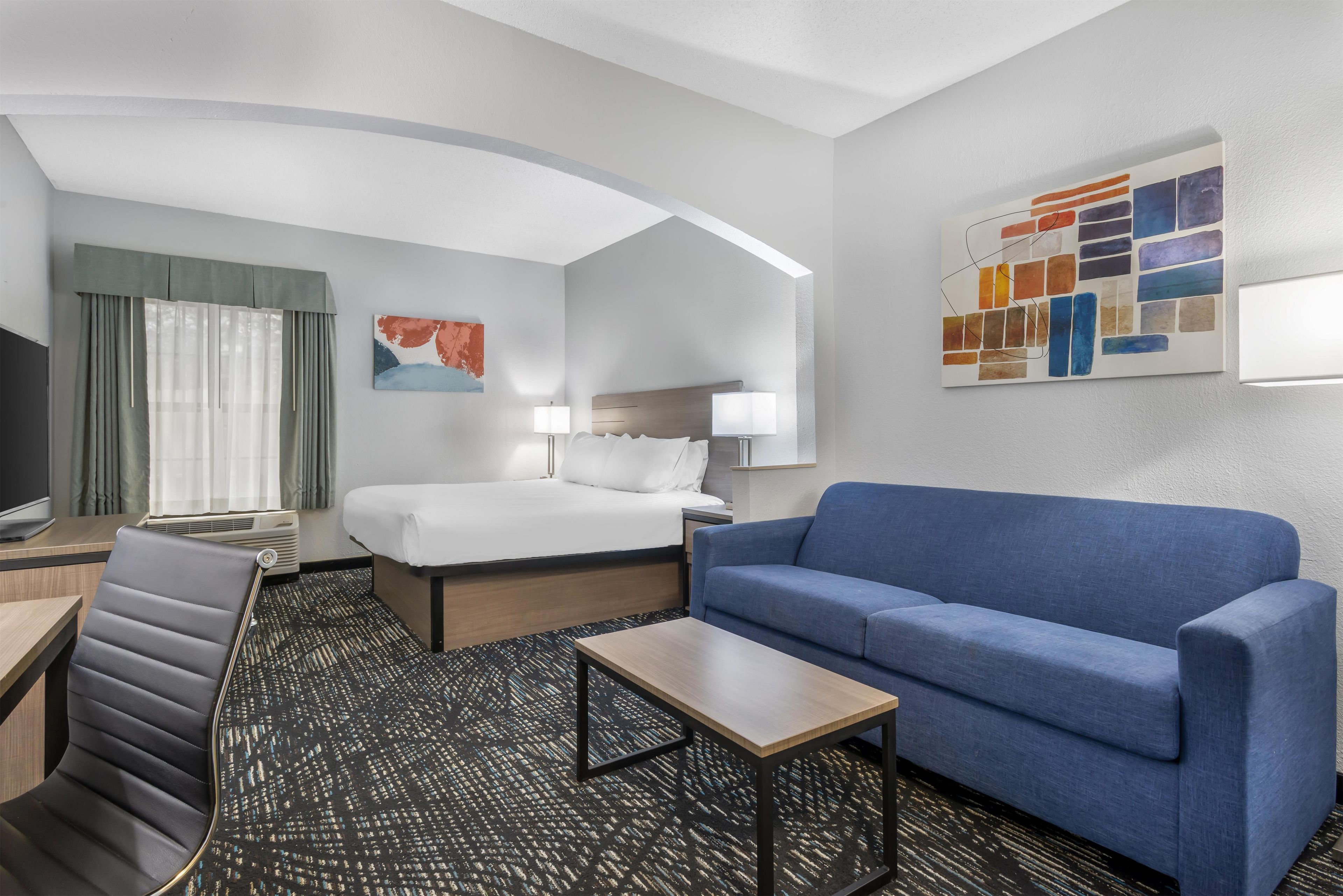 A hotel room with a bed, a blue sofa, a desk with a chair, a TV, and artwork on the walls.