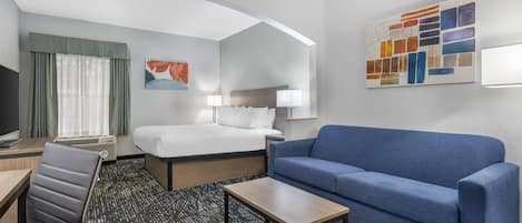 A hotel room with a bed, a blue sofa, a desk with a chair, a TV, and artwork on the walls.