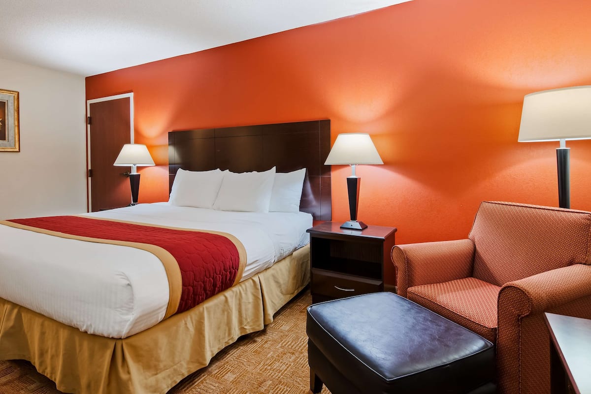A hotel room with a large bed, two bedside tables with lamps, a red armchair, and a small ottoman.
