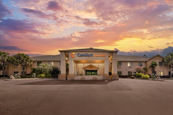 Ein Hotel Eingang mit einem Schild, auf dem 'Comfort Suites' steht, vor einem Sonnenuntergangshimmel.