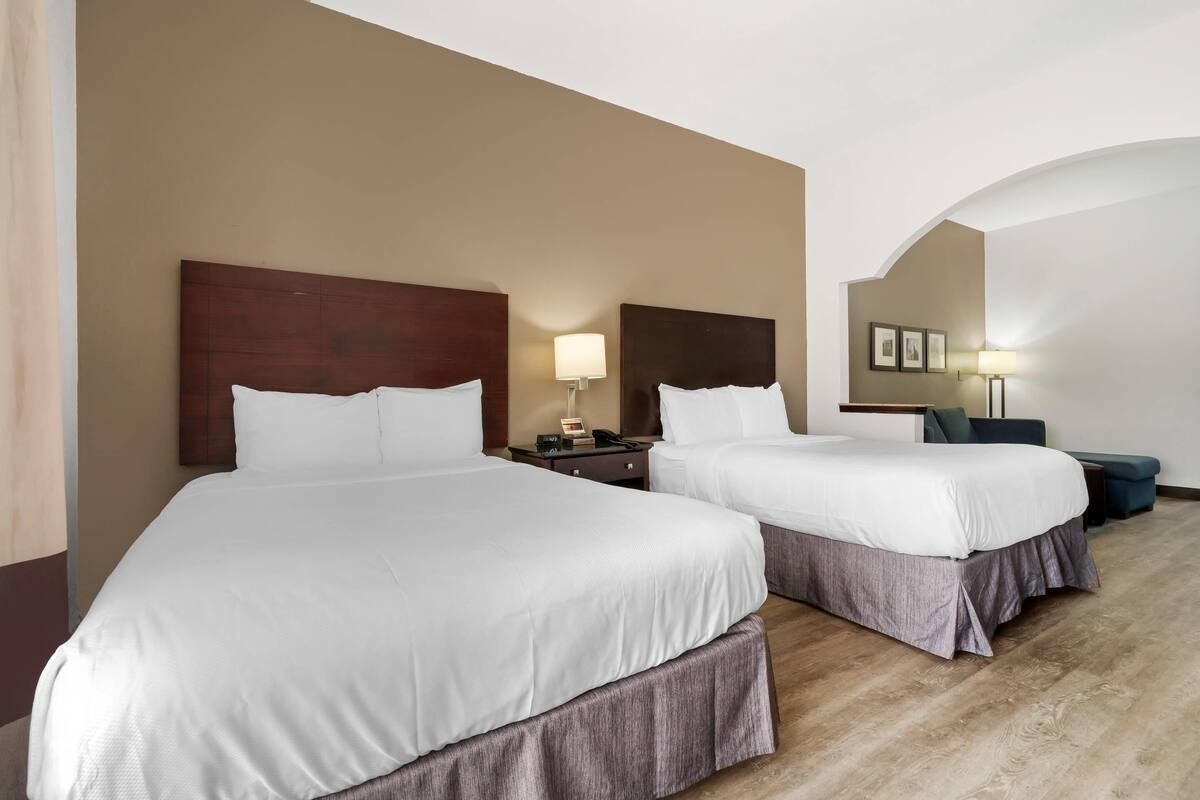 Ein Hotelzimmer mit zwei Betten, einem hölzernen Kopfteil, einem Nachttisch mit Lampe und einer kleinen Sitzecke mit Sessel und Tisch.