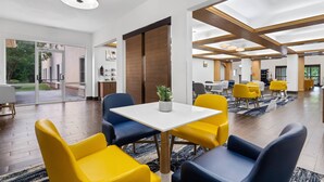 Eine moderne Hotelhalle mit einer zentralen Sitzecke, ausgestattet mit gelben und blauen Stühlen, einem weißen Tisch mit Blumentopf und einer großen Holztür.