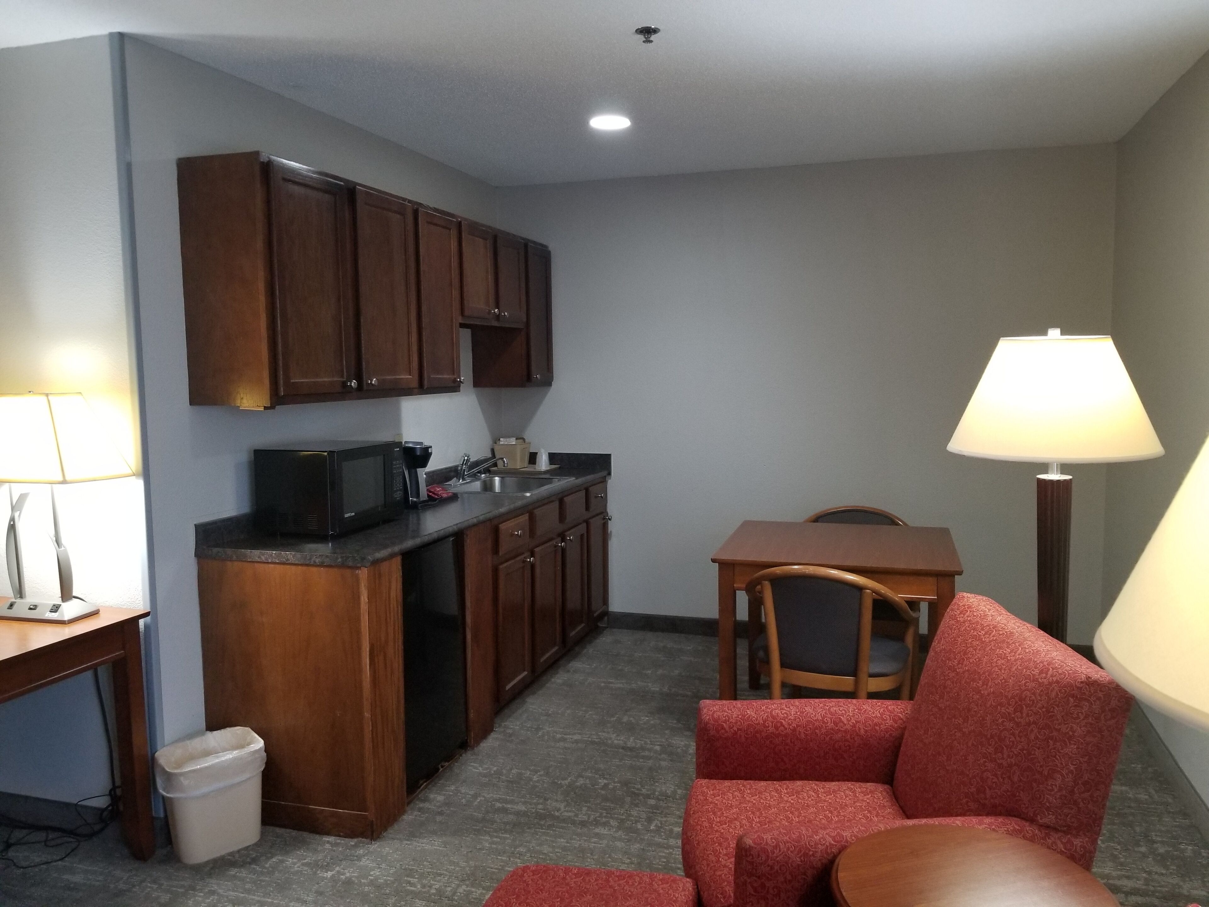 A compact kitchen with wooden cabinets, a microwave, and a sink. There's a small dining area with a table and a red chair. A trash bin is visible near the kitchen.