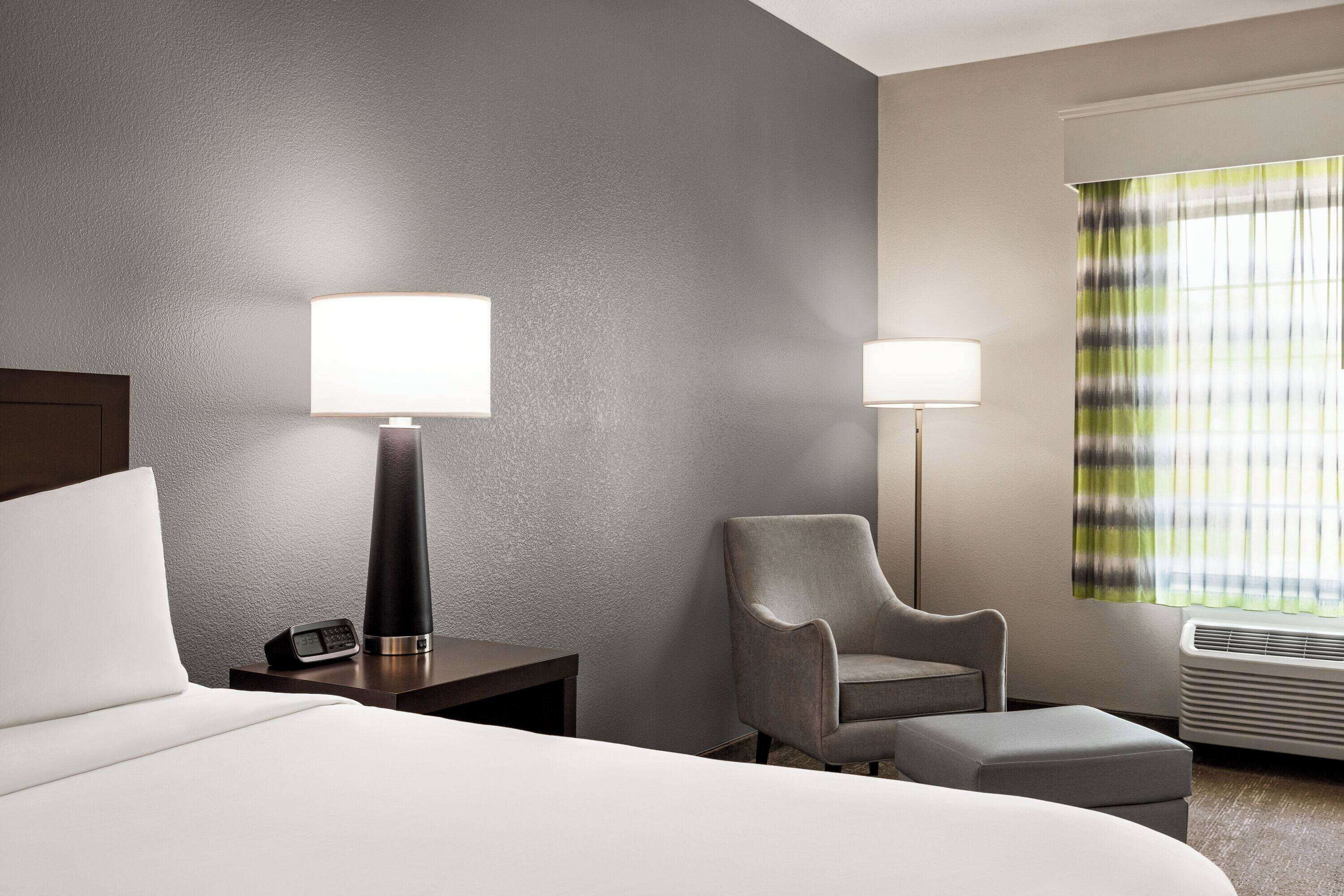 Une chambre d’hôtel avec un lit, une chaise, une table de chevet avec une lampe et une fenêtre avec des rideaux.