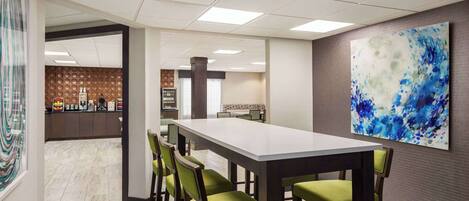 A conference room with a long table and green chairs, a large abstract painting on the wall, and a kitchen area in the background.