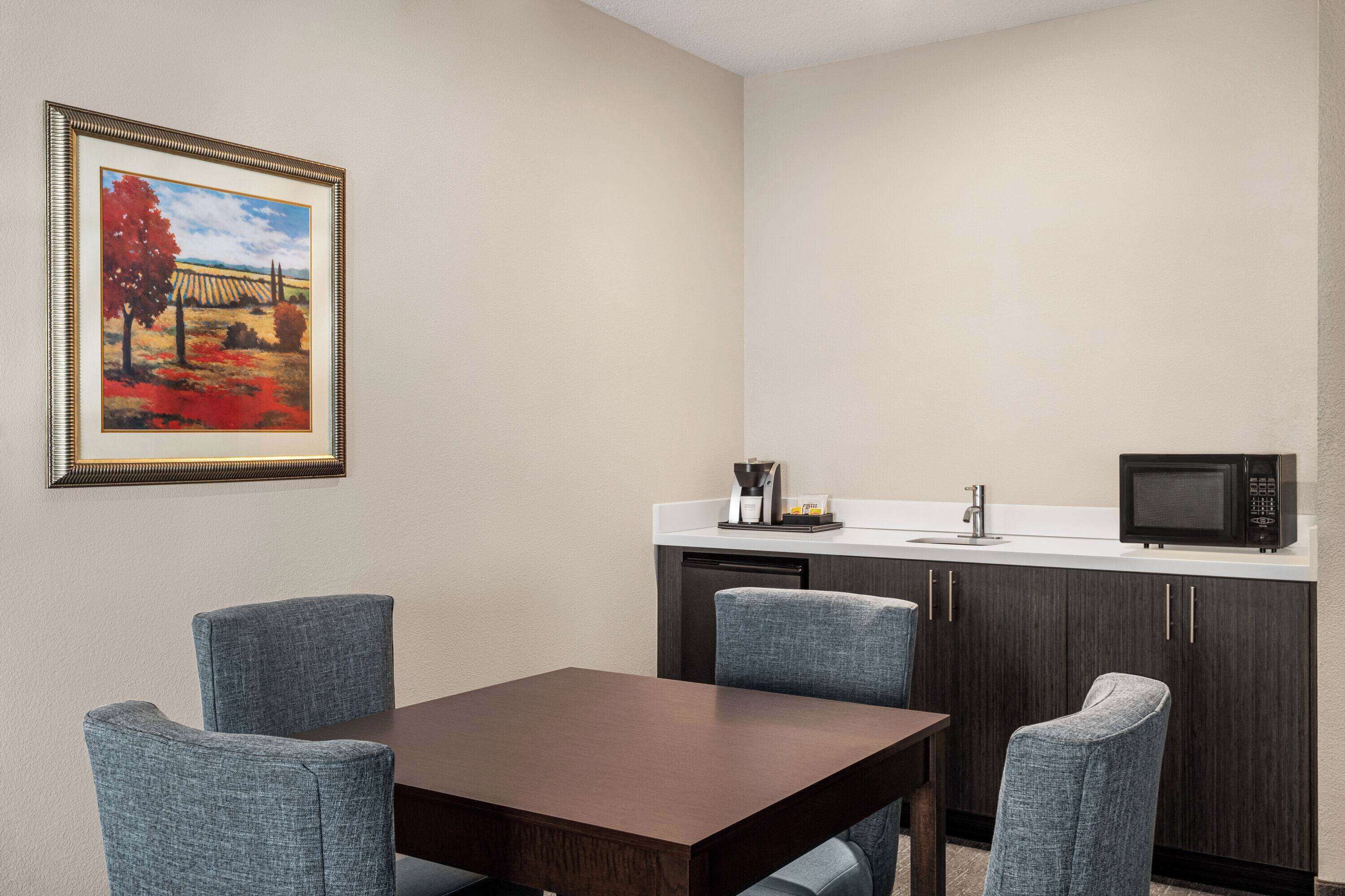 A small, modern room with a wooden table and four blue chairs, a microwave, a coffee maker, and a framed painting on the wall.