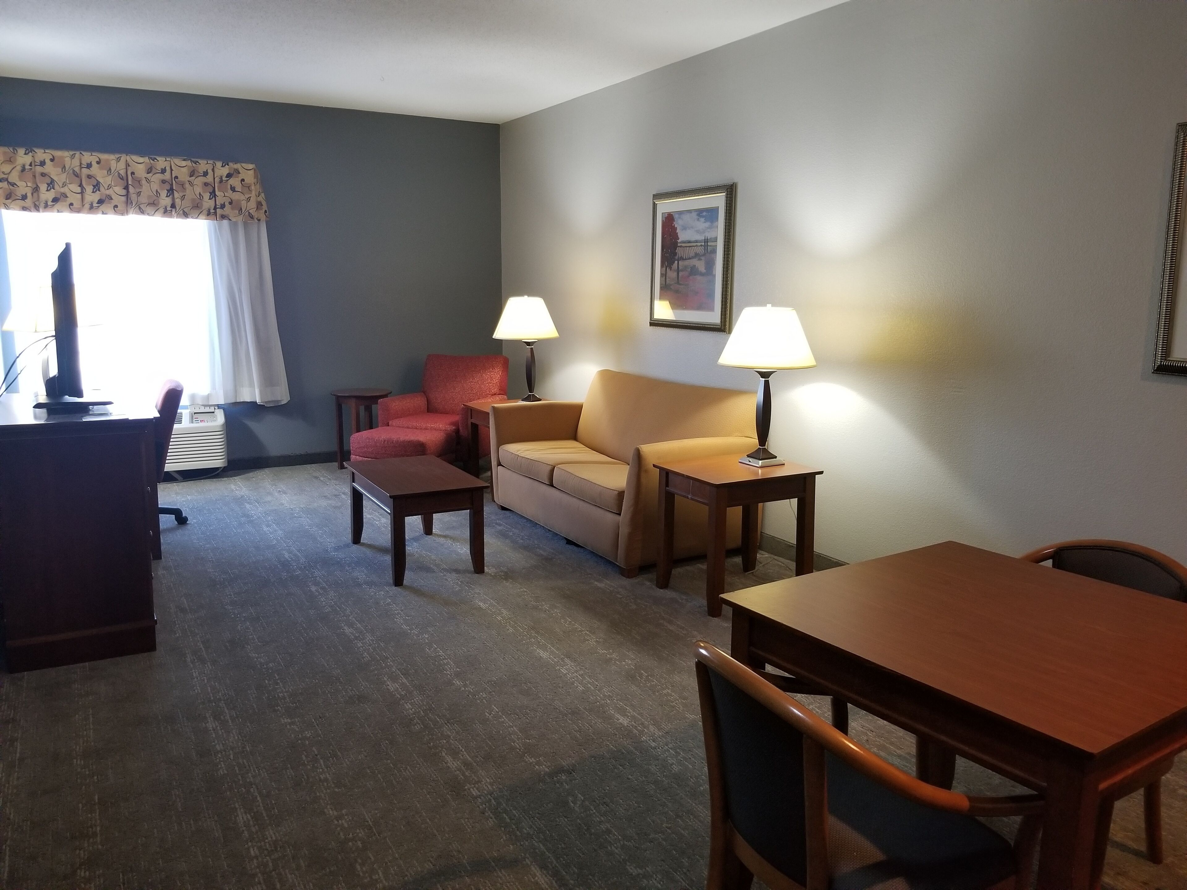Una habitación de hotel con un escritorio, un sillón rojo, un sofá beige, dos lámparas y un cuadro en la pared.