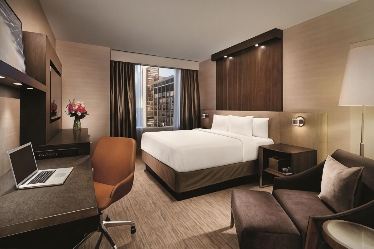 Habitación de hotel con cama, escritorio, laptop, silla, mesita de noche y lámpara.