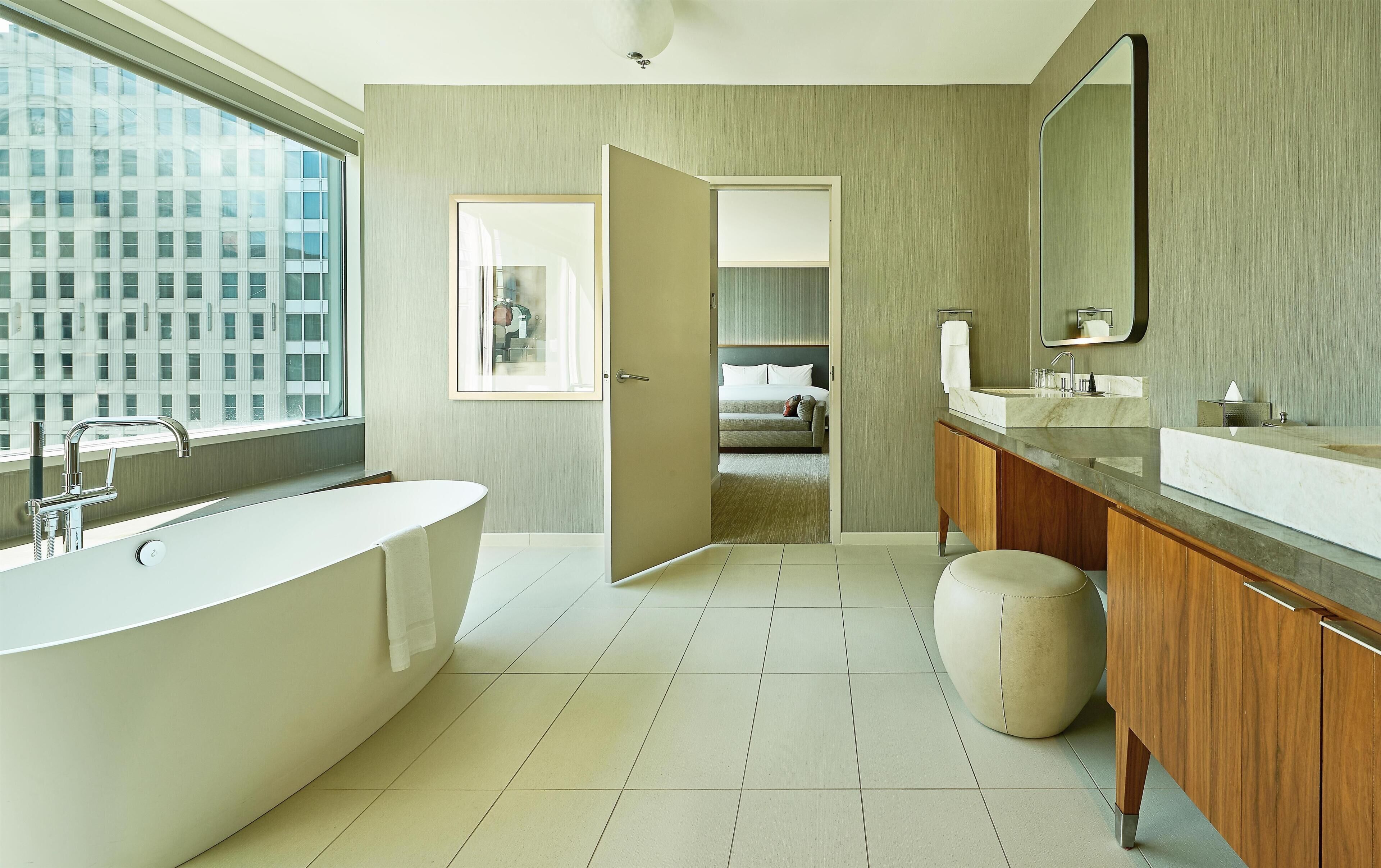 A modern bathroom with a freestanding bathtub, a vanity with a mirror, and a view of a bedroom through an open door.