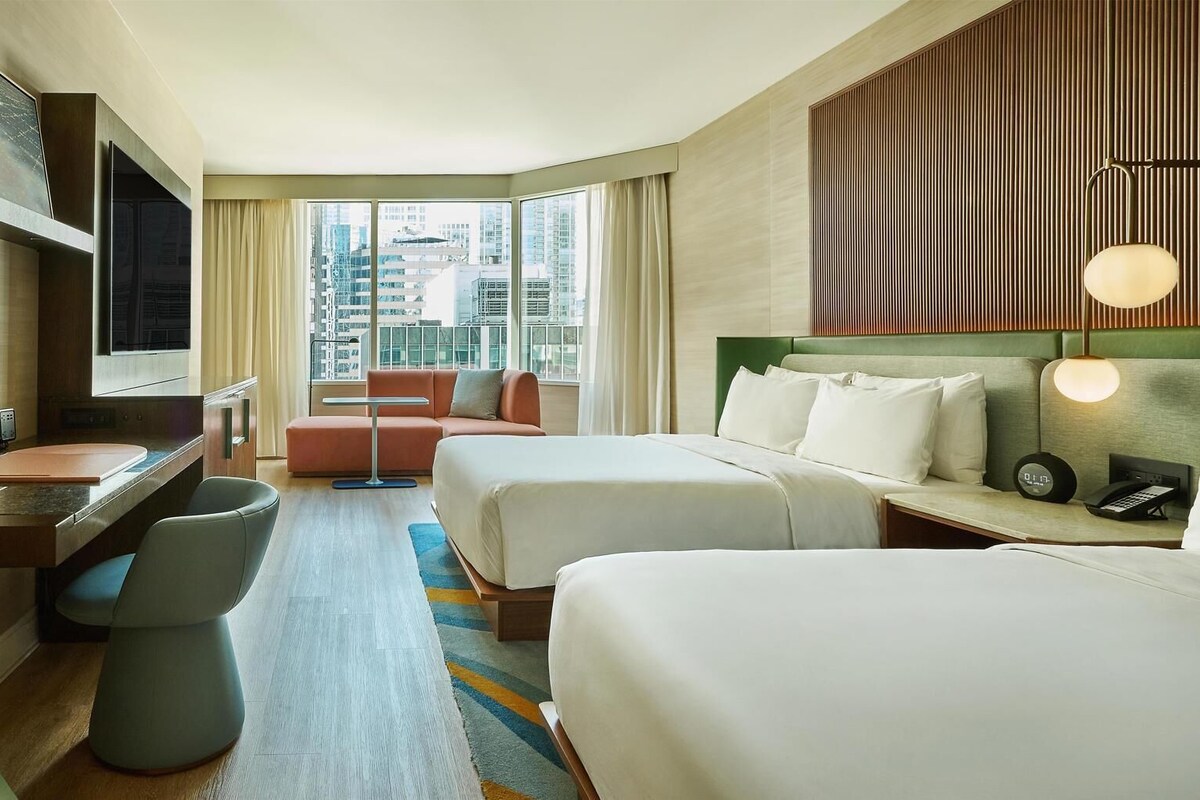 Habitación de hotel con escritorio, dos camas, sofá, televisión y vista a la ciudad.