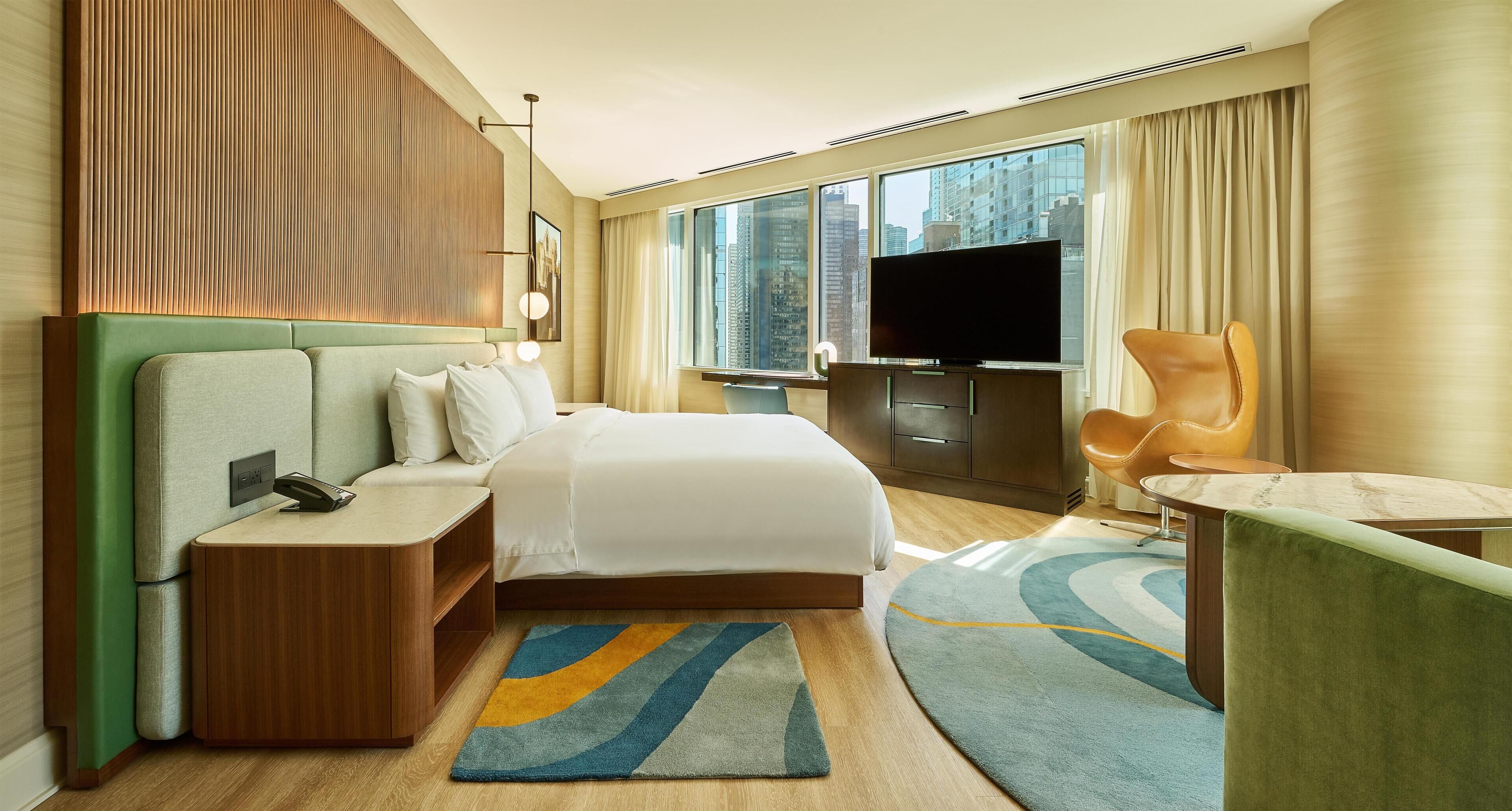 Habitación de hotel con cama, mesita de noche, televisor, zona de estar y vistas a la ciudad.