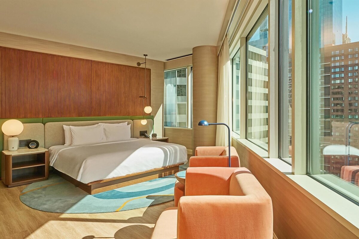 Una habitación de hotel moderna con una cama grande, una zona de estar y vistas a la ciudad a través de amplios ventanales.