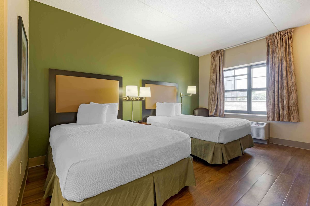 A hotel room with two beds, a large window with curtains, and a wall-mounted air conditioning unit.