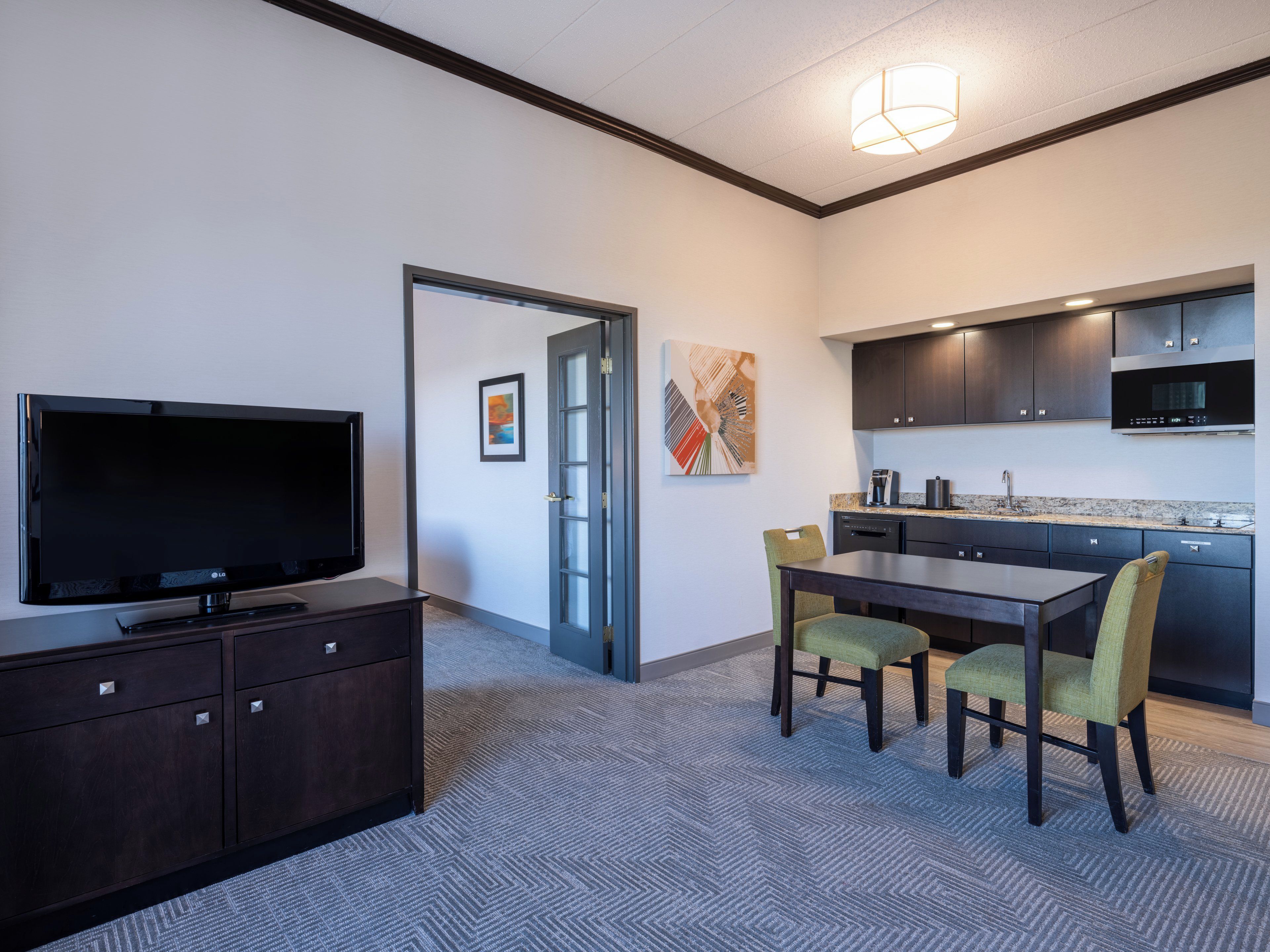 A modern hotel room with a flat-screen TV, a dining area with a table and chairs, a kitchen with dark cabinets and a microwave, and a door leading to another room.