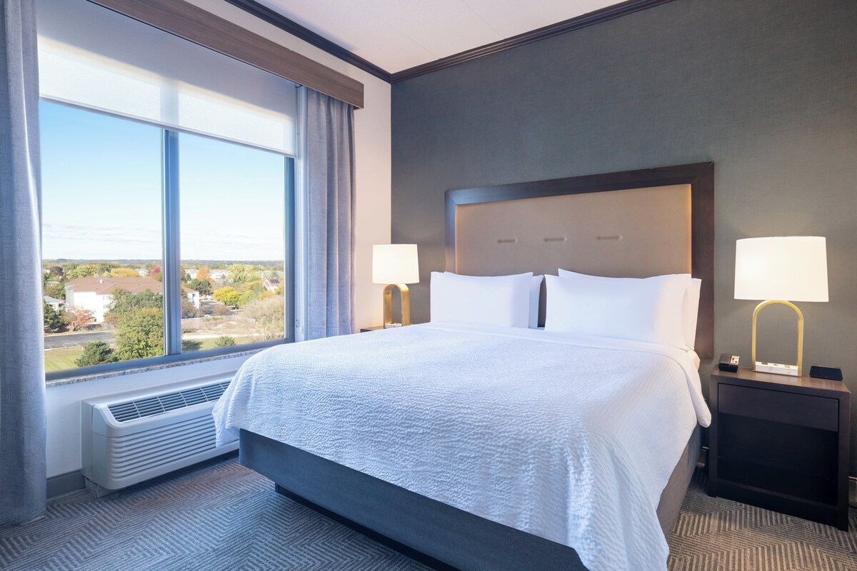 A hotel room with a large bed, two bedside tables with lamps, a window with curtains, and a view of the outside.