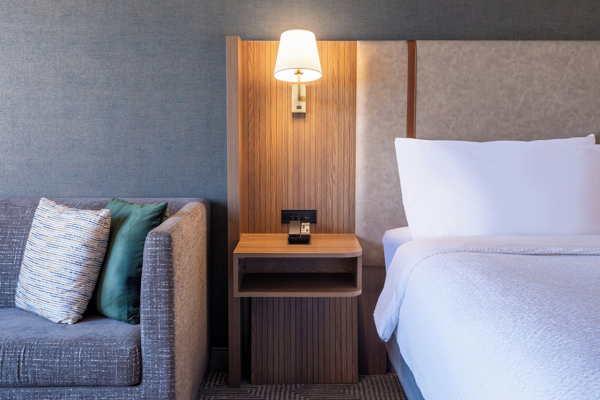 A hotel room with a grey sofa, a wooden nightstand with a lamp, and a bed with white bedding.