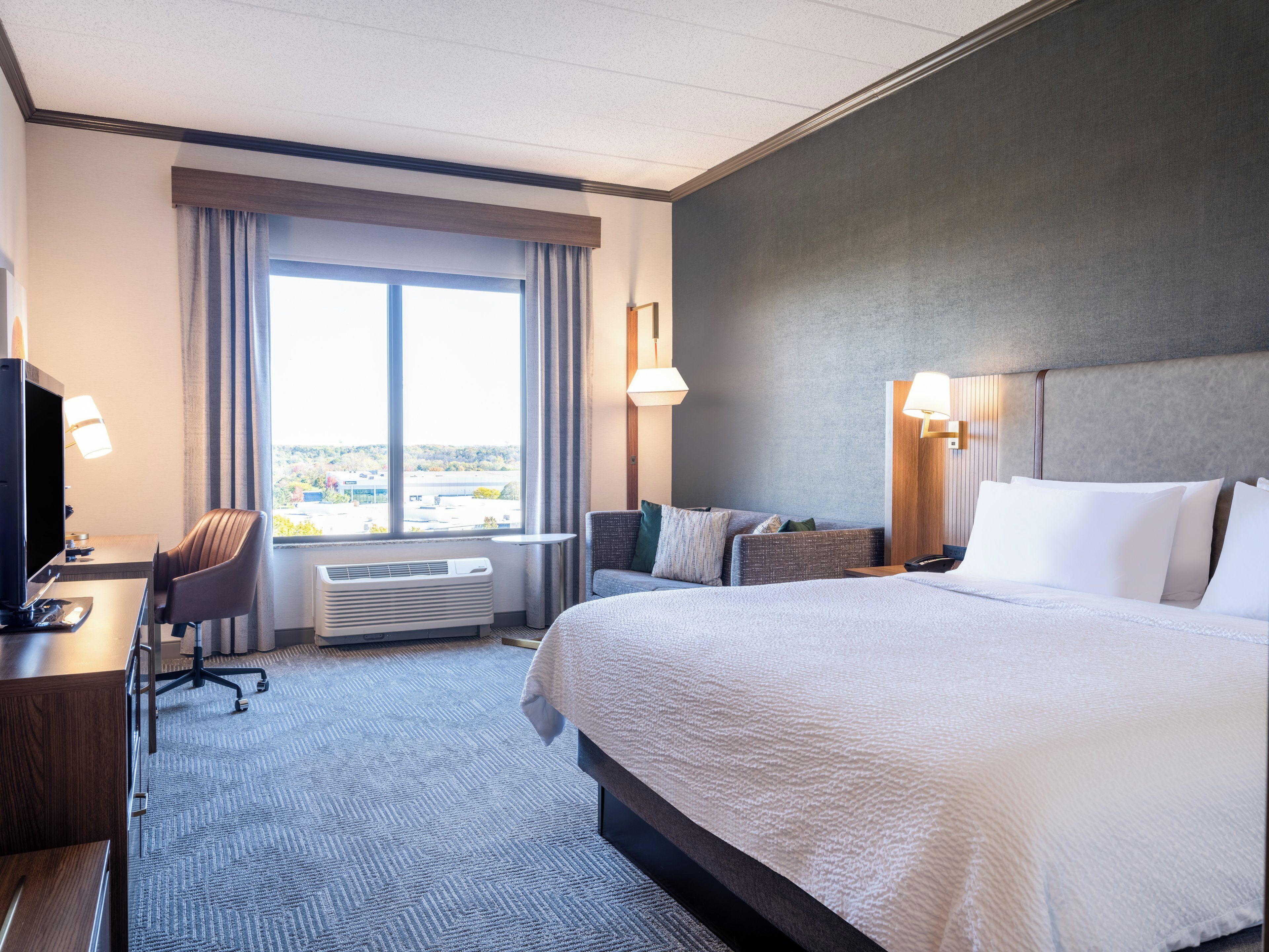 Habitación de hotel con una cama grande, un escritorio con computadora, una silla y un ventanal con cortinas.