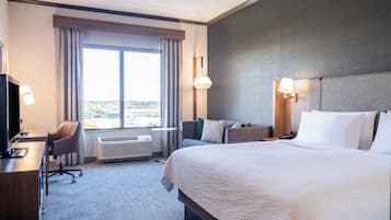 Habitación de hotel con una cama grande, un escritorio con computadora, una silla y un ventanal con cortinas.