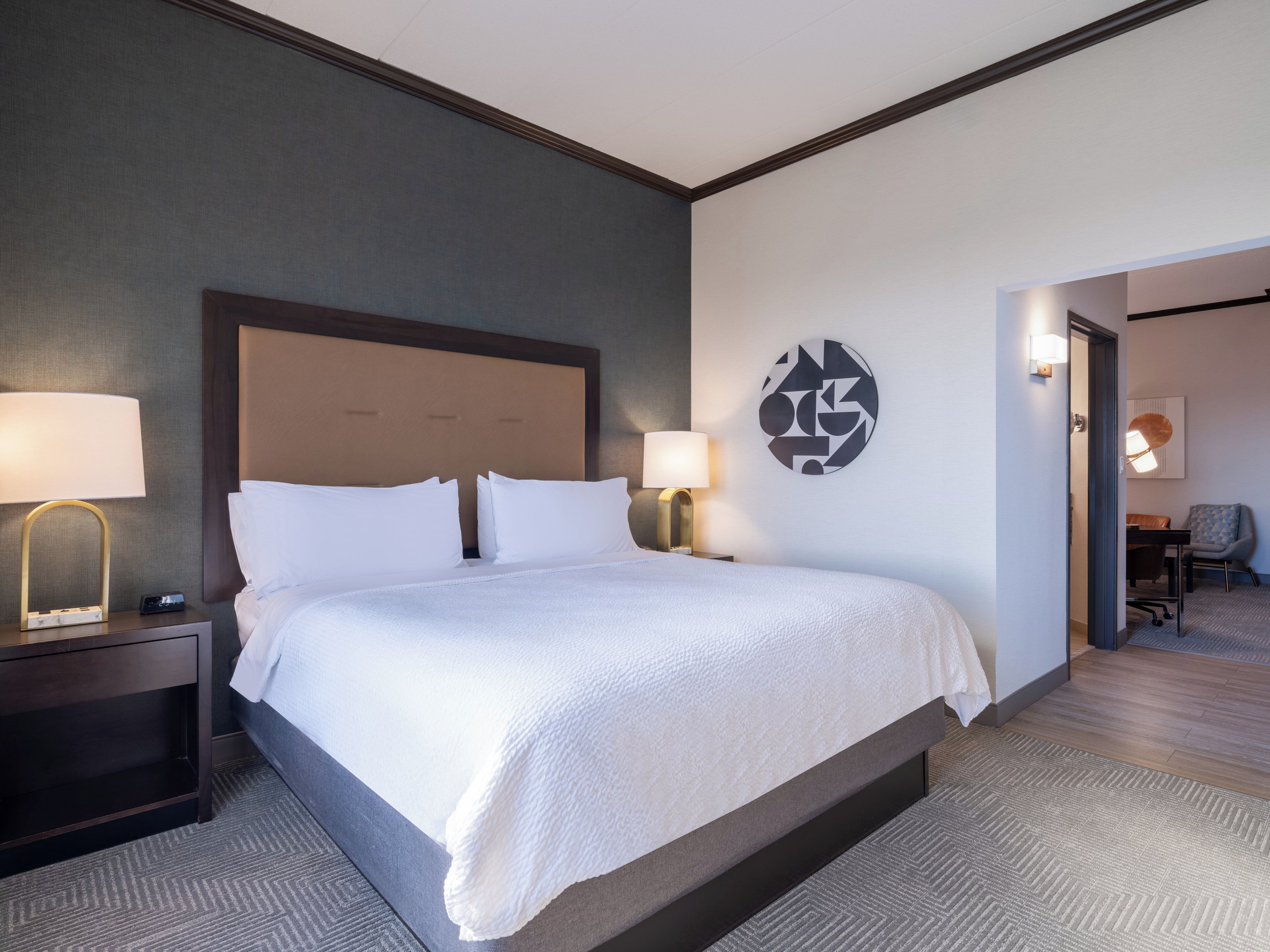 Hotelkamer met een groot bed, twee nachtkastjes met lampen, een kunstwerk aan de muur en uitzicht op een andere kamer.