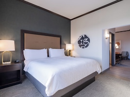 Una habitación de hotel con una cama grande, dos mesitas de noche con lámparas, una obra de arte en la pared y vista a otra habitación.