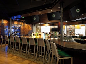 A bar area with a long counter, multiple stools, a television screen, and a variety of bottles and glasses. - Holiday Inn Chicago Northwest-Elgin by IHG (Elgin)